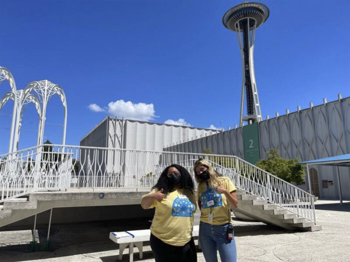 Two camp staffers stand in the PacSci courtyard with the Space Needle and arches in the background.