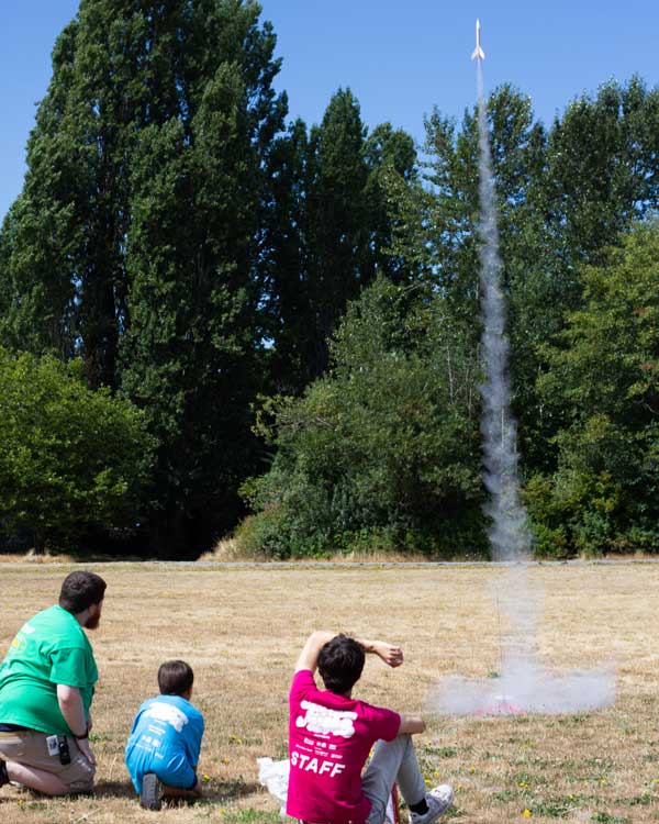 Two camp staff and a camper watch a small rocket in the air
